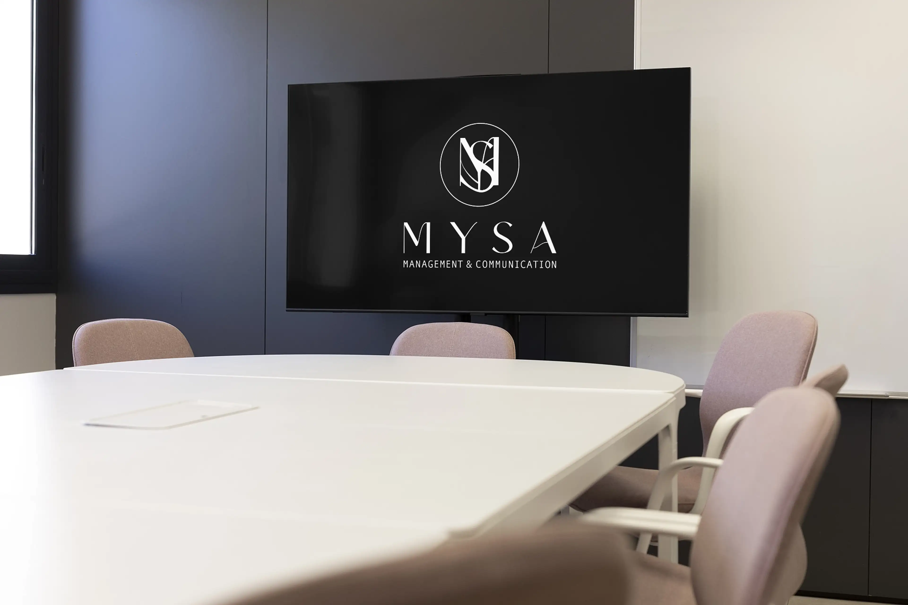 The MYSA logo on a wall-mounted screen in a modern meeting room with blush chairs and a white table